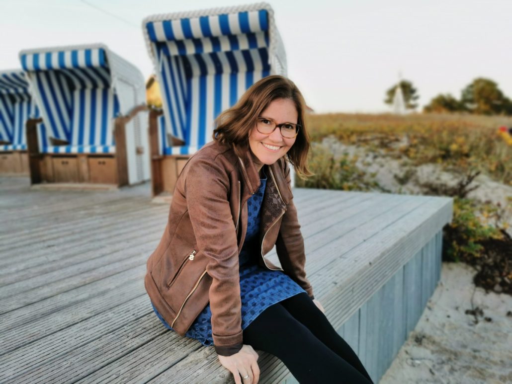 Auf diesem Bild siehst du Kristin Palm. Sie ist Gesundheitscoach. Sie sitzt vor weißen Strandkörben. Kristin trägt eine Brille und hat hellbraune kinnlange Haare. Sie lächelt und ist innerlich zufrieden. Sie ist in einem schmerzfreien Zustand und genießt den Moment.