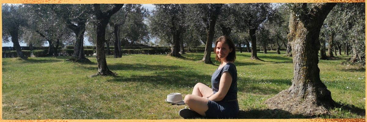 Auf diesem Bild sitzt Kristin Palm- Gesundheitscoach für Frauen auf einer großen Wiese unter sehr alten Olivenbäumen. Sie hat gerade meditiert und schau nun tiefenentspannt in die Kamera. Es ist Sommer und sie strahlt von innen heraus.