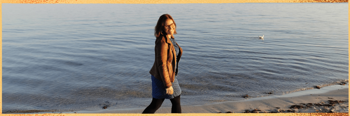 Auf diesem Bild siehst du Kristin Palm. Sie macht einen Strandspaziergang bei Sonnenschein im Herbst. Sie lächelt und ist glücklich. Sie zeigt sich Sorgen- und Schmerzfrei und schaut zufrieden aus. Sie ist im Einklang mit ihrem Körper und Geist. Sie Schaut über ihre linke Schulter in die Kamera und im Hintergrund ist die Ostsee zu sehen.