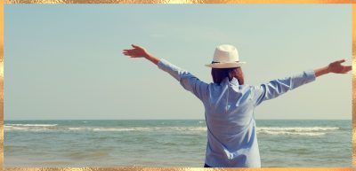 Hier steht eine Frau, vermutlich Kristin Palm-für Körper, Geist und Seele am Strand und breitet die Arme jubelnd gen Himmel aus. Ihr Blick ist Richtung Wasser gerichtet. Sie trägt ein blaues Hemd und einen weißen Strohhut.