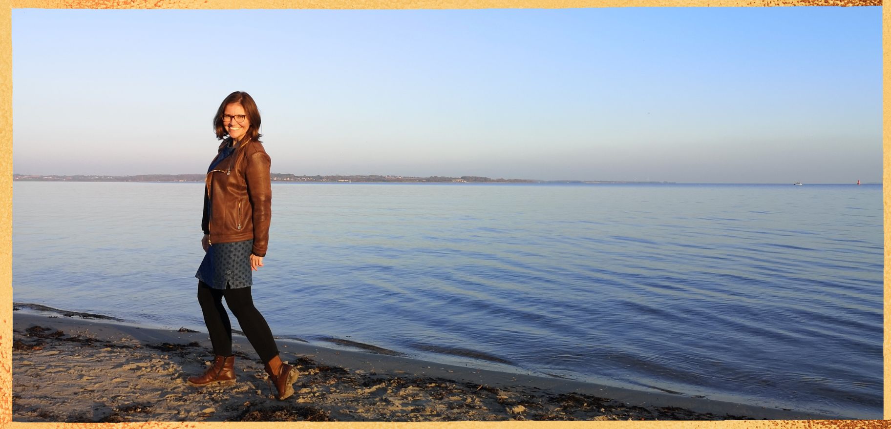 Hier siehst du Kristin Palm- für Körper, Geist und Seele am Strand, im Hintergrund ist die Ostsee. Kristin lächelt und schuat über ihre linke Schulter in die Kamera. es scheint die Sonne.