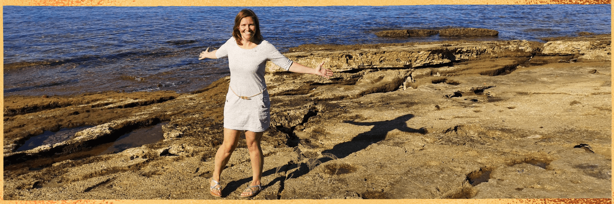 Hier siehst du Kristin Palm. Sie dreht sich etwas im Kreis und ist sehr fröhlich. Sie hat ein weißes Kleid an, strahlt in die Kamera, und breitet beide Arme entspannt zur Seite aus. Im Hintergrund siehst du schroffe Felsenformationen und das Meer.