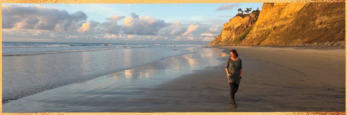 Hier ist Kristin Palm zu sehen, die über ihre langjährige Arbeit als Physiotherapeutin, ihre Leidenschaft für Yoga und Meditation sowie ihre heutige Tätigkeit als Spiritual Life Coach spricht und ihre Mission beschreibt, Menschen zu mehr Kraft, Balance und innerer Stärke zu begleiten.“ Hier steht Kristin am Strand von San Diego bei Sonnenuntergang.