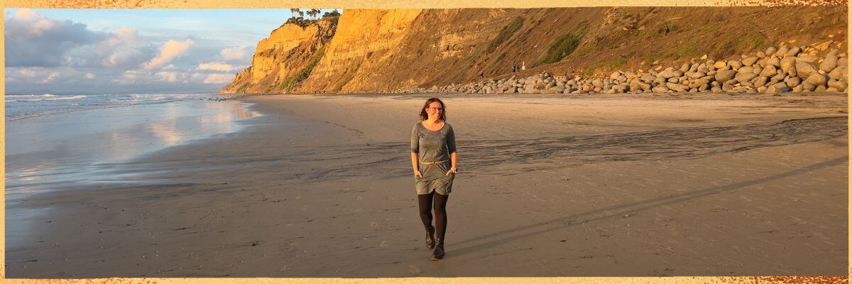 Hier ist Kristin Palm zu sehen, die über ihre langjährige Arbeit als Physiotherapeutin, ihre Leidenschaft für Yoga und Meditation sowie ihre heutige Tätigkeit als Spiritual Life Coach spricht und ihre Mission beschreibt, Menschen zu mehr Kraft, Balance und innerer Stärke zu begleiten.“ Hier steht Kristin am Strand von San Diego bei Sonnenuntergang.